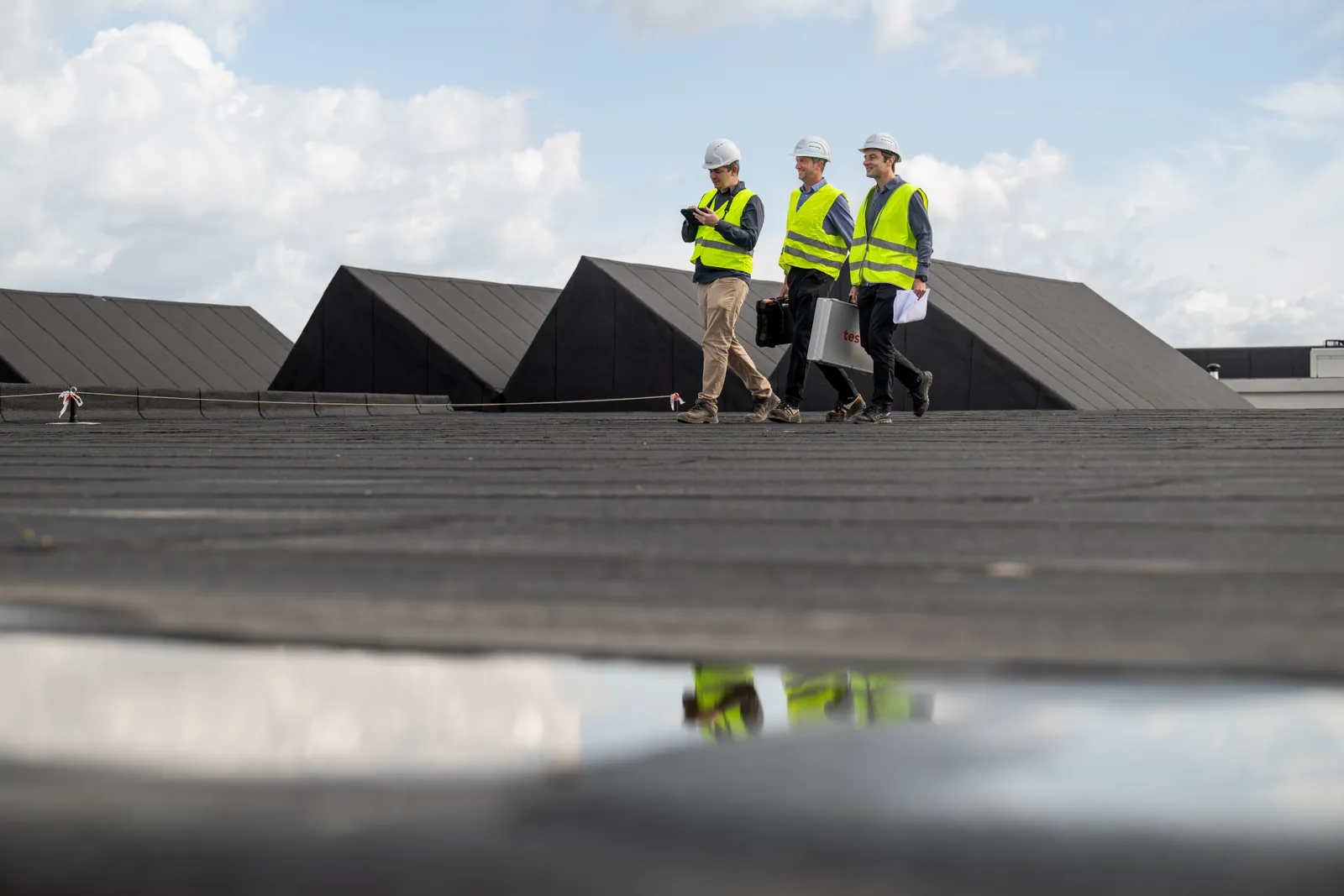 Drie ingenieurs in fluohesje en witte helm lopen over het platte dak langs de zwarte sheddaken, weerspiegeld in een plas op de voorgrond.