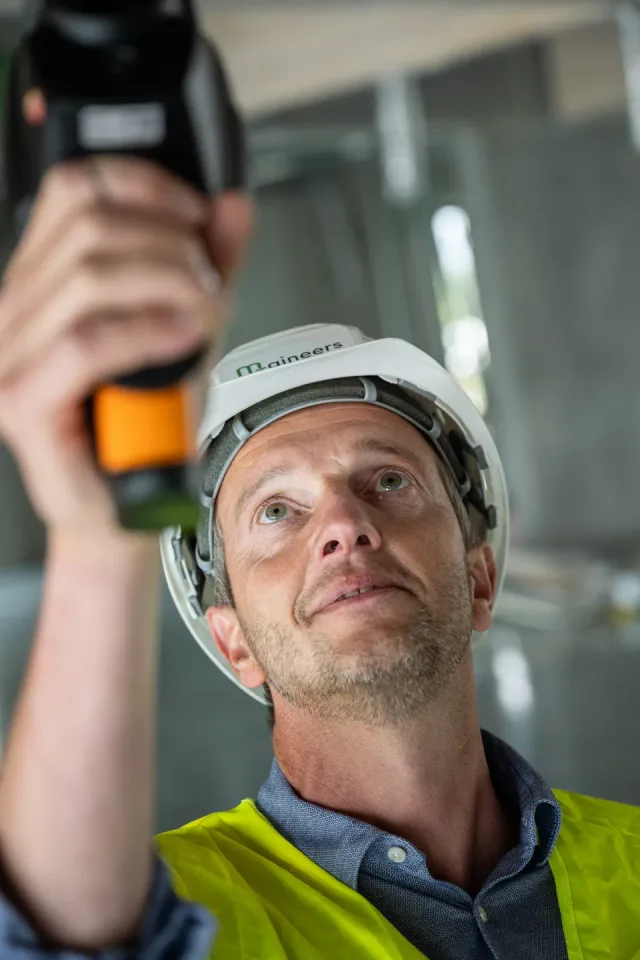Close-up van een ingenieur met M-gineers-helm die opkijkt naar het meettoestel dat hij boven zich houdt, in een ruwbouw.