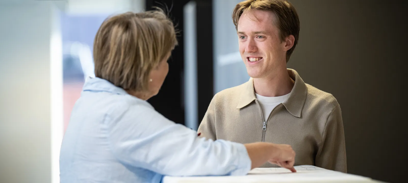 Twee collega's aan de balie: een vrouw in lichtblauw wijst iets aan op een document, de man in beige luistert glimlachend mee.