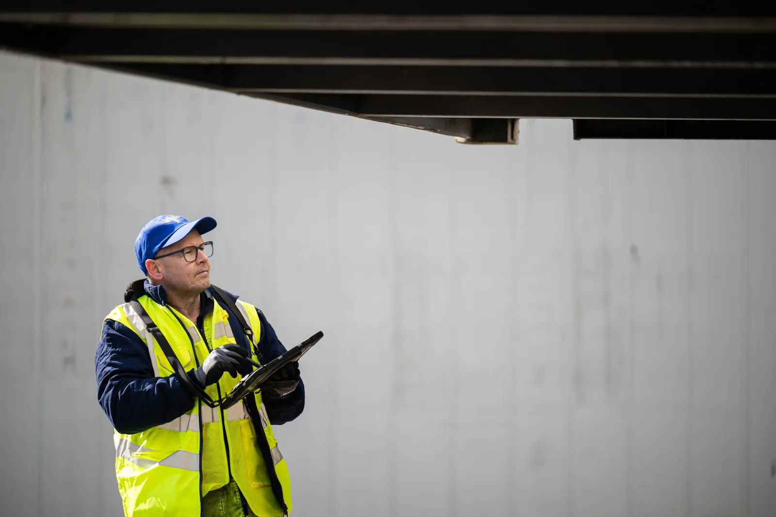 Een medewerker in fluohesje, bril en blauwe Thornton-pet kijkt op van de tablet in de hand naar de container die boven het hoofd uitsteekt.