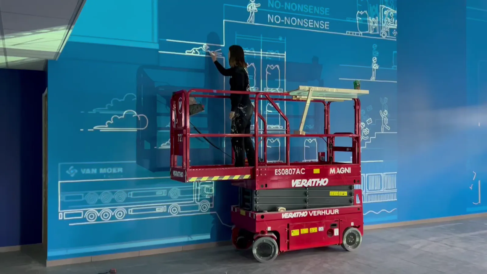 De muurschildering in de maak: een schilder op een hoogtewerker trekt de witte lijn op de blauwe Academy-muur.