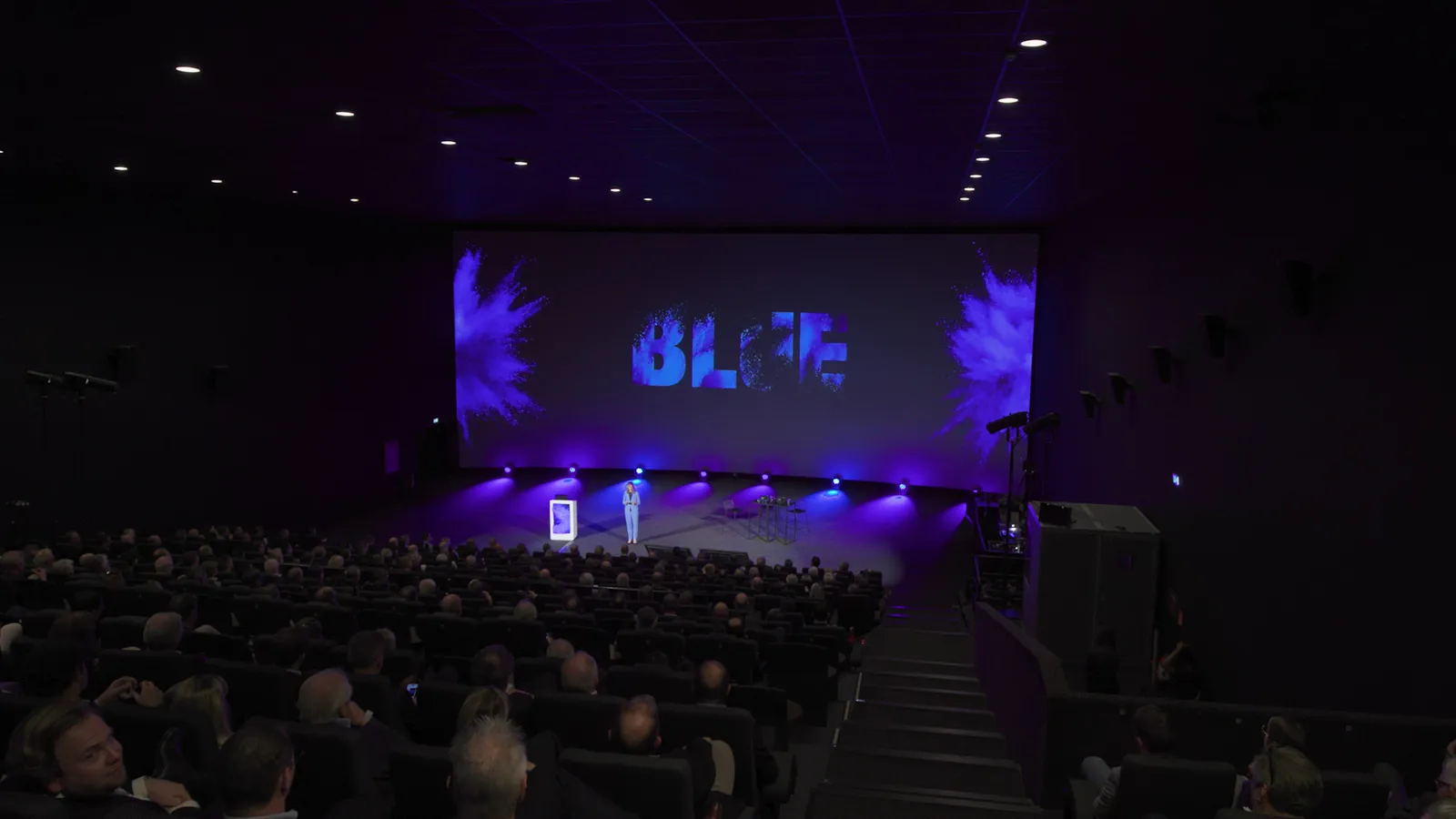 Beeld uit de Blue-aftermovie: de volle cinemazaal van het klantenevent, de presentatrice op het podium onder het BLUE-scherm.